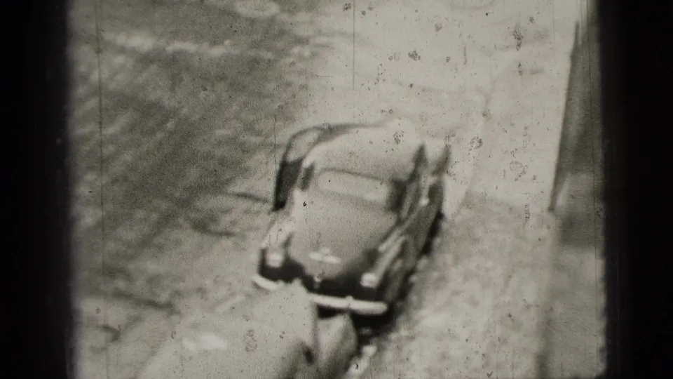 High-angle 1956 New York City home-movie frame showing the parked mid-century car after the man moves partly out of view.