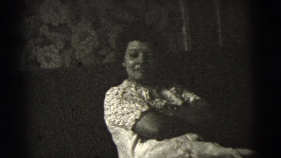 Seated woman indoors in 1956 home-movie footage, arms crossed and facing the camera with a faint smirk.