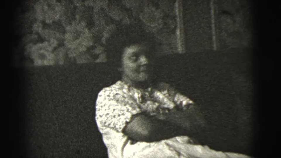 Indoor 1956 home-movie frame of a woman seated with crossed arms, turned slightly away amid dim domestic surroundings.