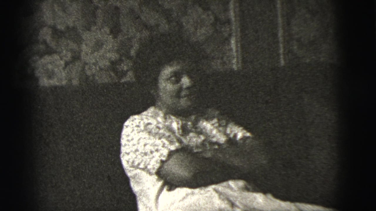 In 1956 New York City home-movie footage, an adult woman sits on a sofa with arms crossed against floral wall covering.