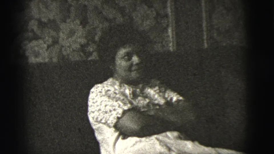 In 1956 New York City home-movie footage, an adult woman sits on a sofa with arms crossed against floral wall covering.