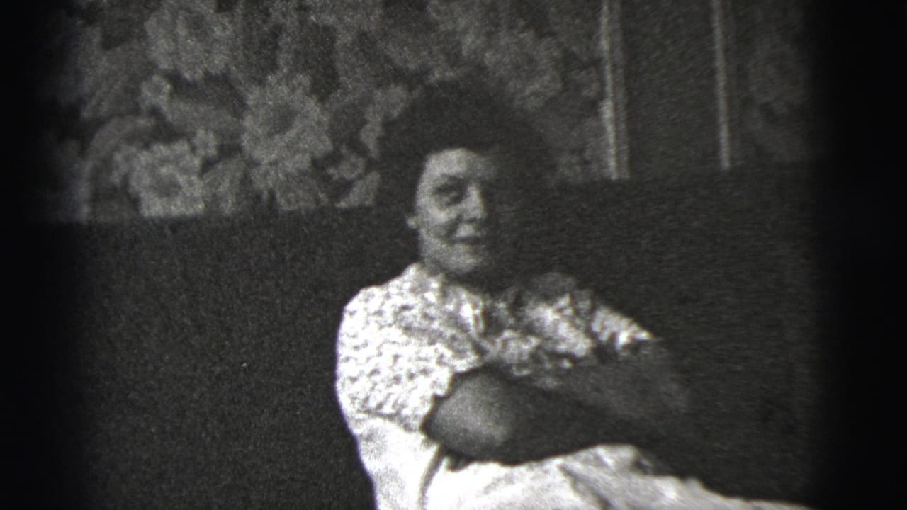 The woman remains seated on the sofa, facing more toward the camera in black-and-white 8mm footage from the private interior.