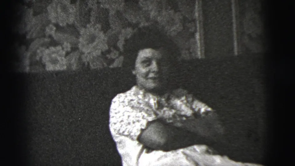 The woman remains seated on the sofa, facing more toward the camera in black-and-white 8mm footage from the private interior.