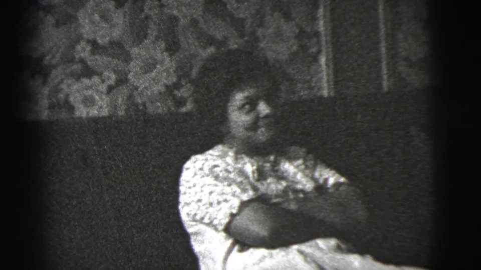 A woman in a patterned blouse sits on a sofa and looks to the side, framed by soft 8mm grain and floral wall covering.