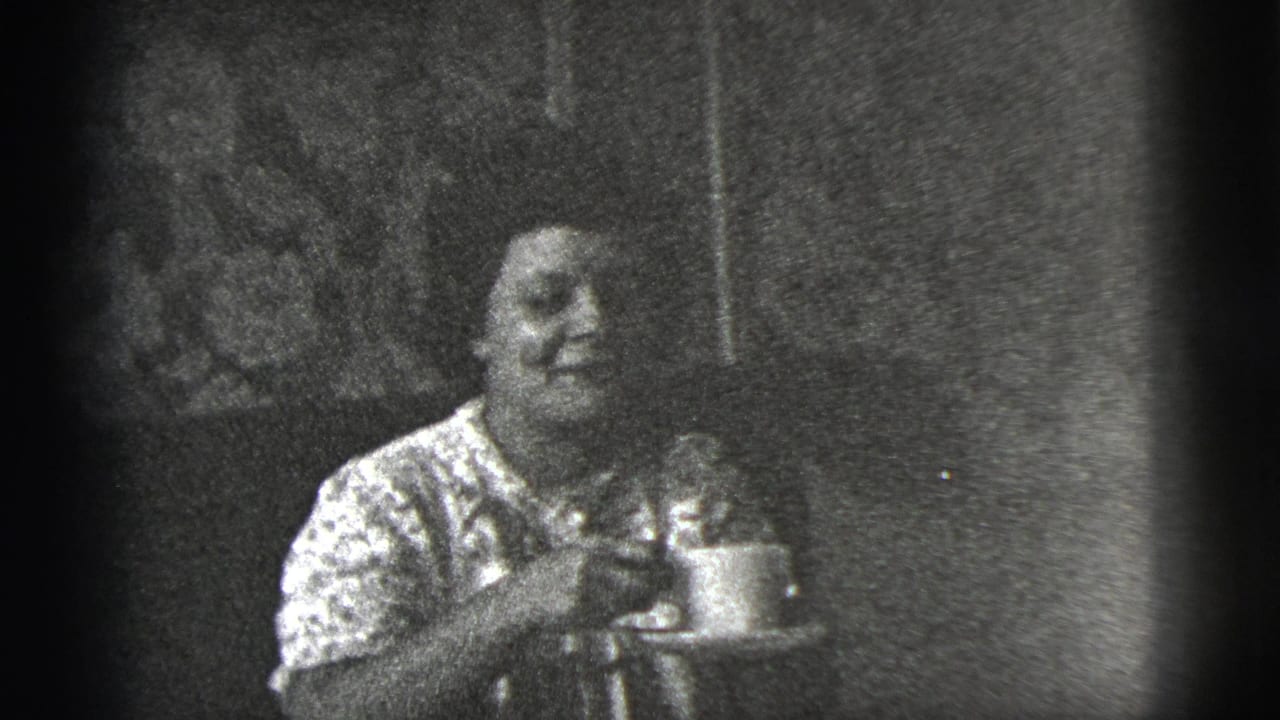 In 1956 New York City home-movie footage, a woman in a patterned blouse sits indoors holding a cup and saucer.