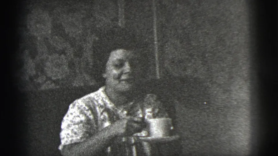 In 1956 New York City home-movie footage, a woman in a patterned blouse sits indoors holding a cup and saucer.