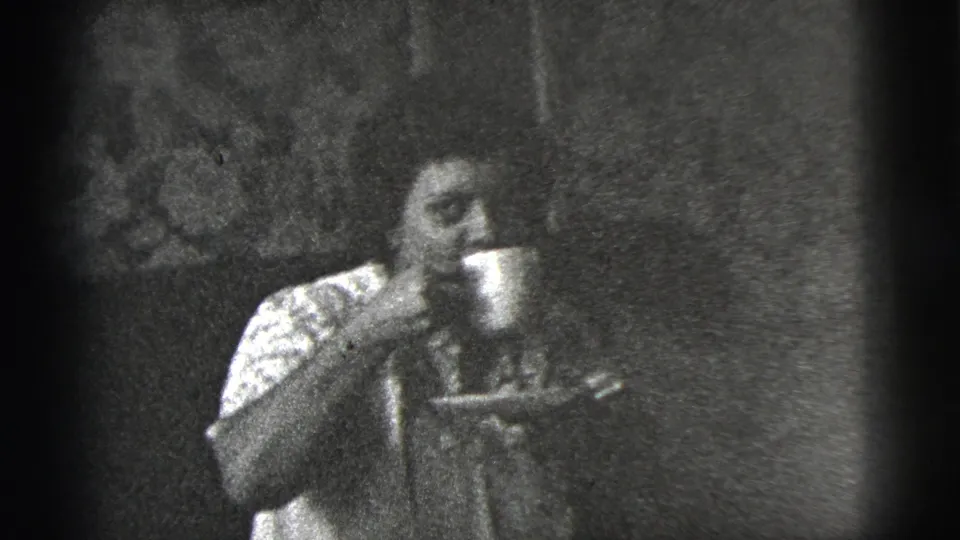 A woman in a patterned blouse raises a cup to drink while seated in a 1956 New York City interior.