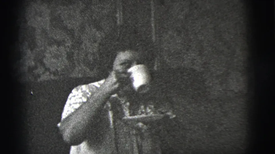 The seated woman drinks from her cup, with the saucer held below, in black-and-white 8mm home-movie footage.