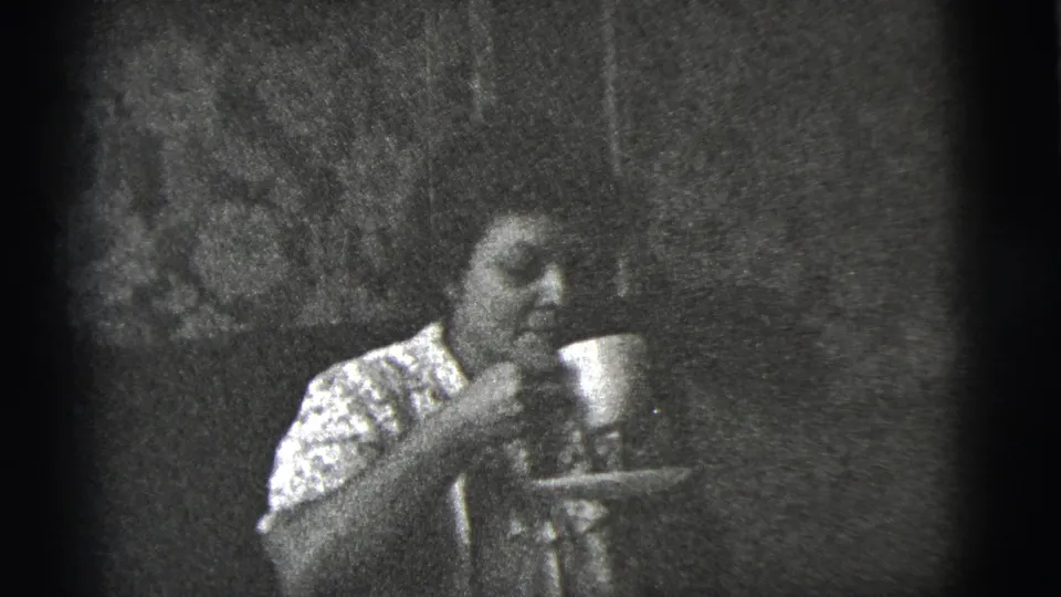 In 1956 New York City home-movie footage, a seated woman in a patterned dress holds a cup and saucer in a dim interior.