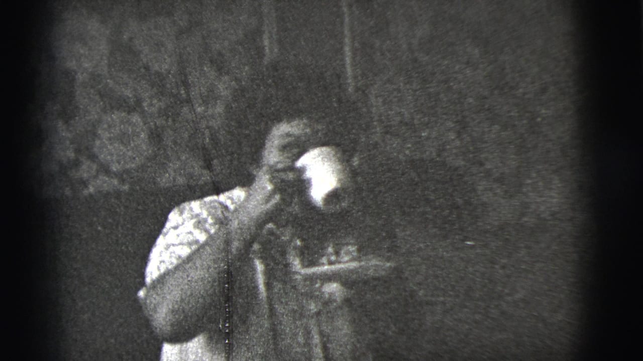 The woman raises a cup to drink while seated indoors, with her face partly obscured by the cup in the 1956 8mm frame.