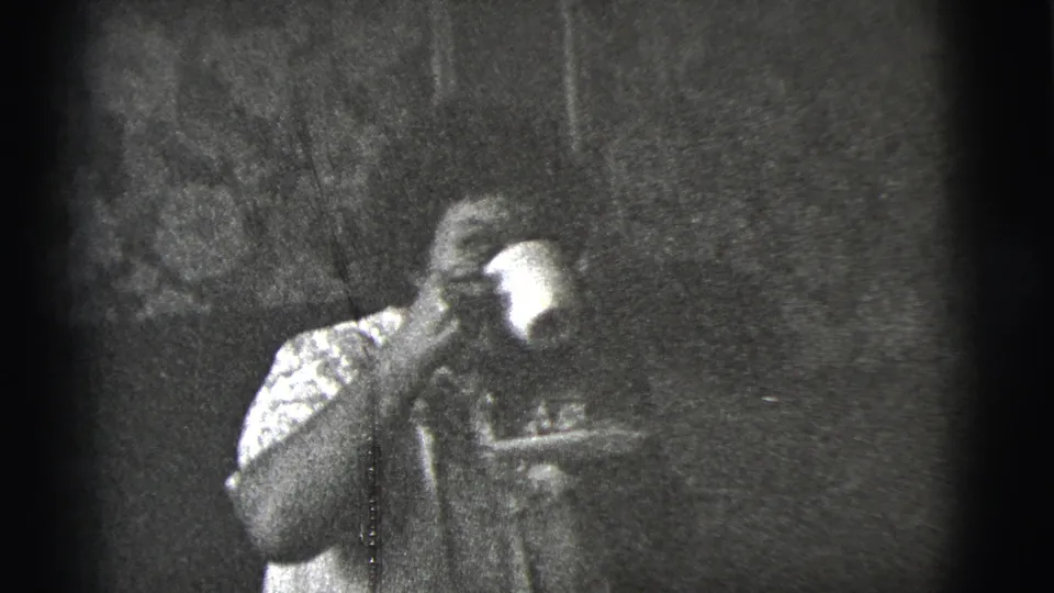 The woman raises a cup to drink while seated indoors, with her face partly obscured by the cup in the 1956 8mm frame.