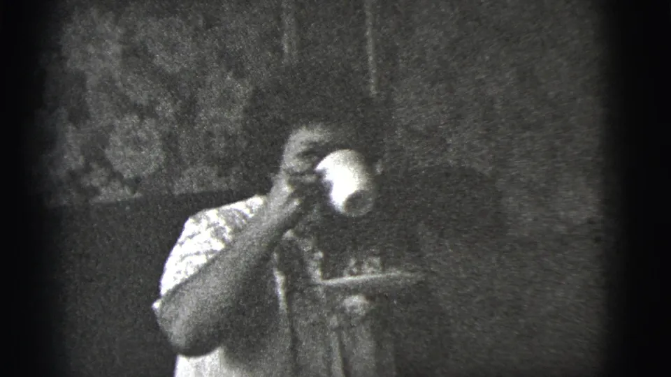 A seated woman sips from a light cup in a dim private interior, captured in grainy black-and-white 8mm footage.
