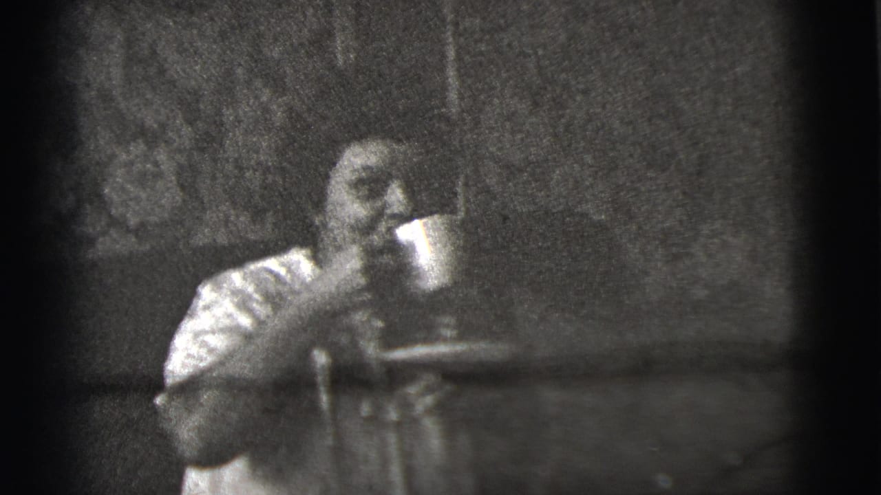 Grainy 1956 8mm frame from New York City showing a seated female figure holding a cup near a dark background.