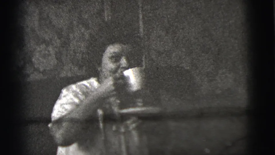 Grainy 1956 8mm frame from New York City showing a seated female figure holding a cup near a dark background.