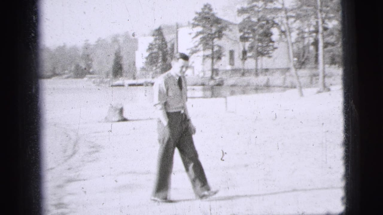 1956 New York City home-movie frame showing a uniformed man walking across a bright park area near trees and a building.