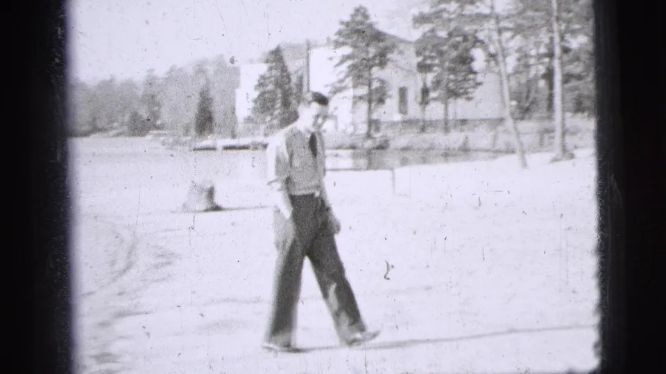 1956 New York City home-movie frame showing a uniformed man walking across a bright park area near trees and a building.