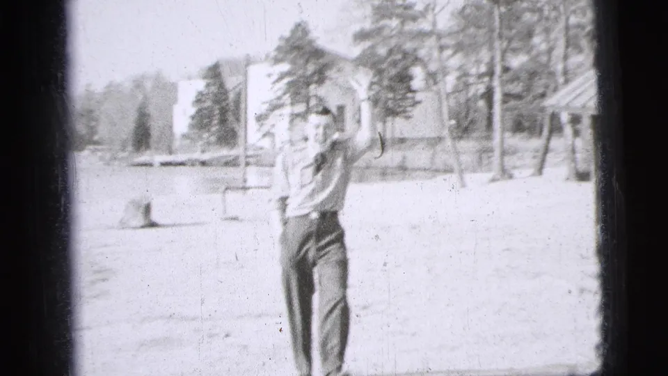 1956 New York City frame of a uniformed man facing the home-movie camera and waving in an open park area.