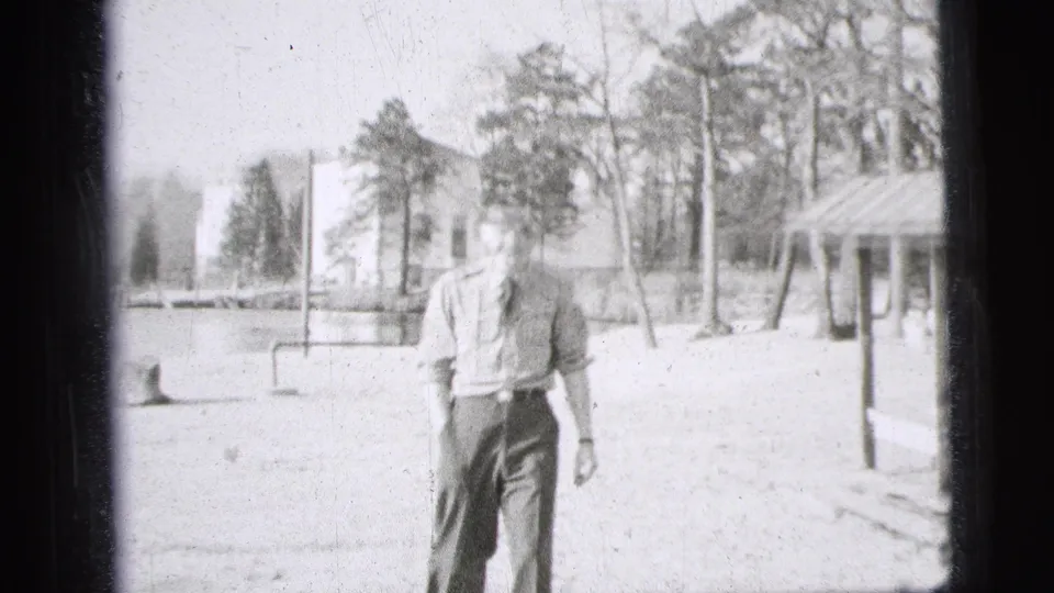 1956 New York City 8mm frame showing a uniformed man standing in a park landscape with trees, a building and a shelter structure.