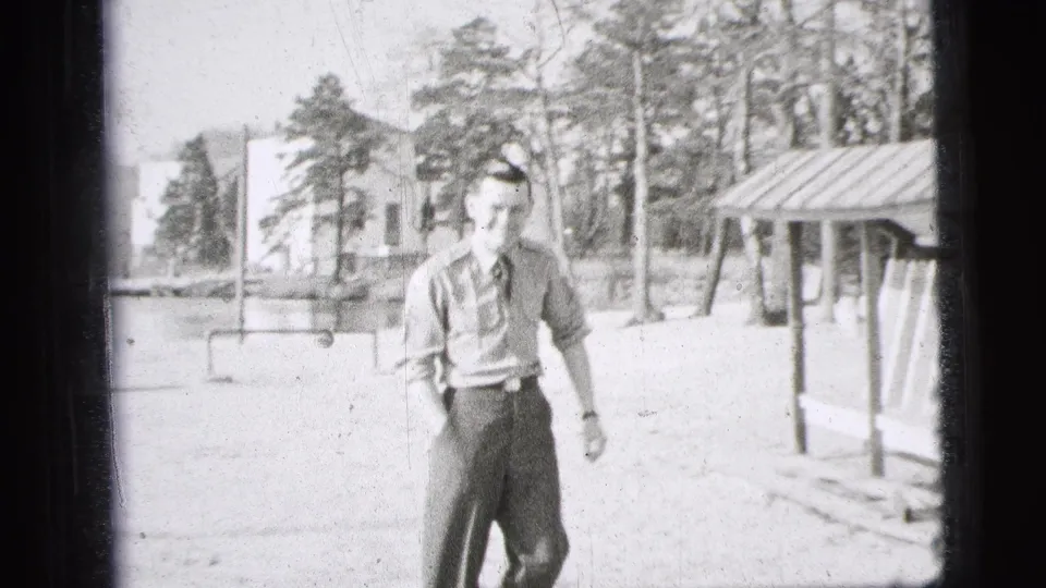 A young man in a short-sleeved military-style uniform walks toward the camera in bright outdoor 1956 New York City home-movie footage.