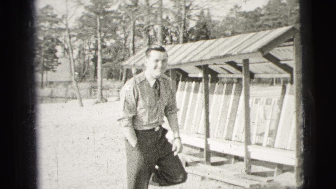 The man in uniform smiles near a wooden shelter, with trees and open ground visible in the 1956 home-movie frame.