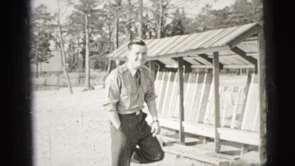 The man in uniform smiles near a wooden shelter, with trees and open ground visible in the 1956 home-movie frame.