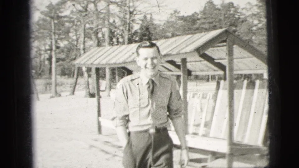 A uniformed man laughs and faces the camera in a bright outdoor setting with a wooden shelter and trees behind him.