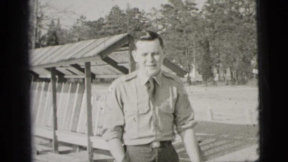 The man poses close to the camera in uniform, one hand in his pocket, with the shelter and tree line behind him.