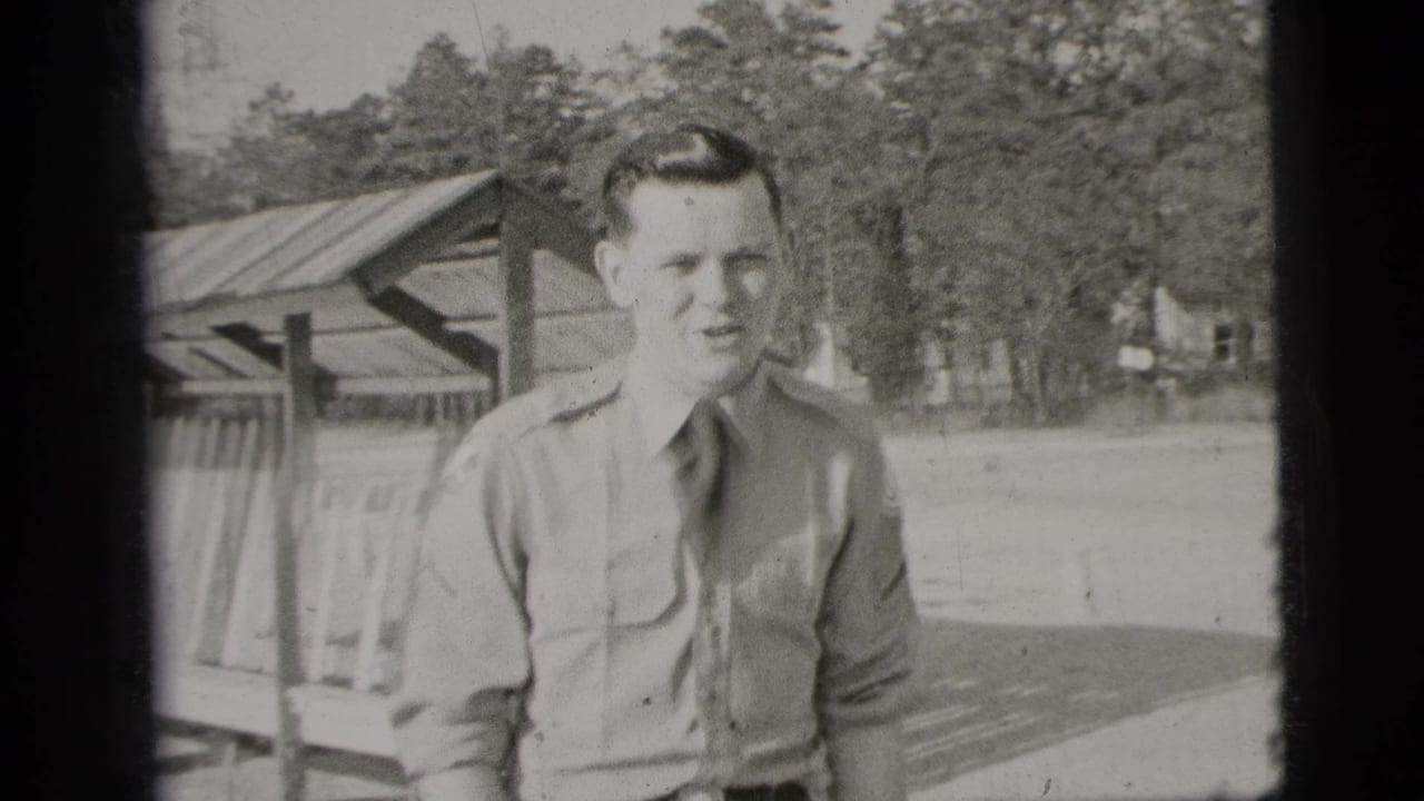 1956 New York home-movie frame showing a young man in a uniform-style shirt standing outdoors with trees and a wooden shelter behind him.