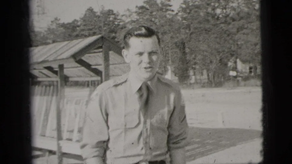 1956 New York home-movie frame showing a young man in a uniform-style shirt standing outdoors with trees and a wooden shelter behind him.