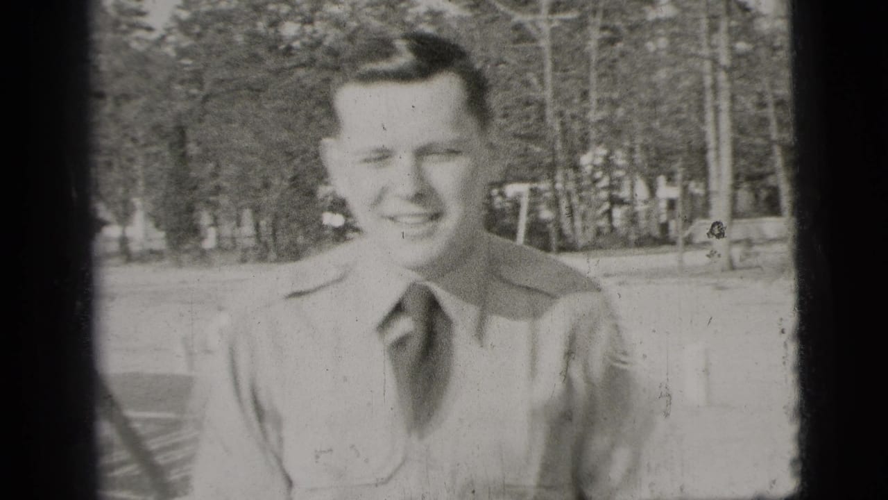 Black-and-white 1956 New York frame of a young man in a uniform-style shirt smiling at the camera with trees in the background.