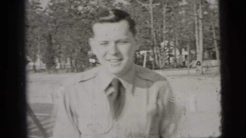 Black-and-white 1956 New York frame of a young man in a uniform-style shirt smiling at the camera with trees in the background.