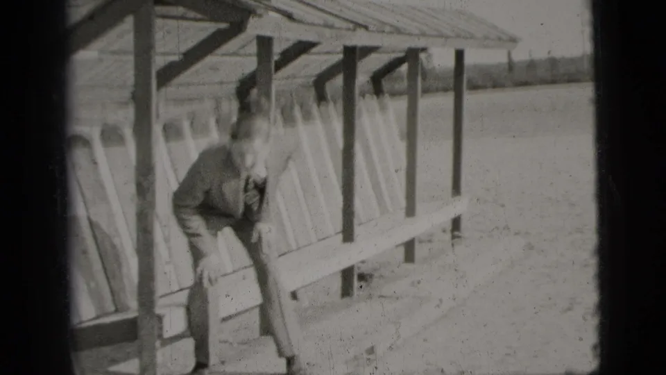 In 1956 New York City home-movie footage, a suited man leans forward on a bench beneath an open shelter near a lake.
