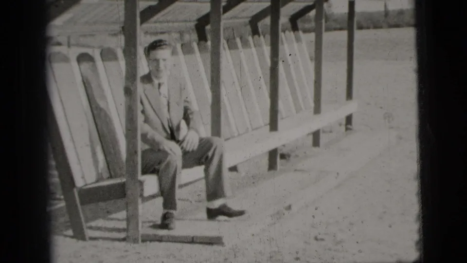 The suited man sits upright on the lakeside bench under the shelter, facing the camera in the 1956 New York City clip.