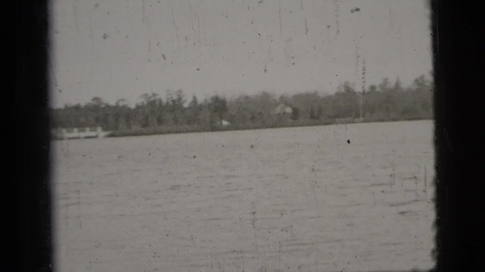 The final frame shifts to the lake, showing calm water, a distant treeline, and shoreline in the same black-and-white home-movie sequence.