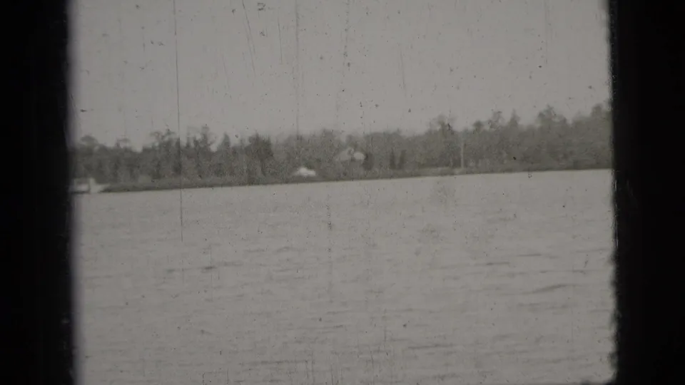 Black-and-white 1956 New York City 8mm frame showing open water, a wooded far shore, and dark vertical film-edge borders.