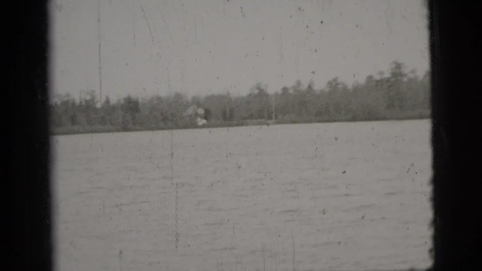 Black-and-white 1956 New York City home-movie frame with open water, a distant wooded shore, and a small light structure across the water.