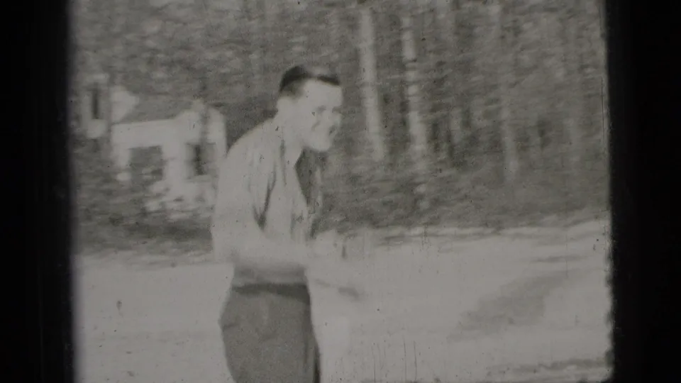 An adult man in a light shirt and trousers moves through a wooded outdoor area in the supplied 1956 New York City footage.