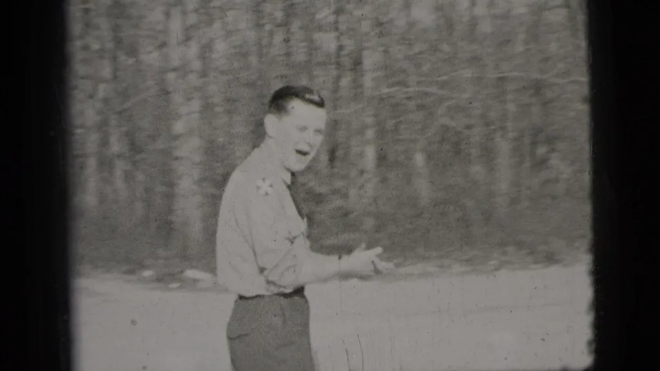 A smiling adult man gestures outdoors near bare trees in black-and-white 1956 New York City home-movie footage.