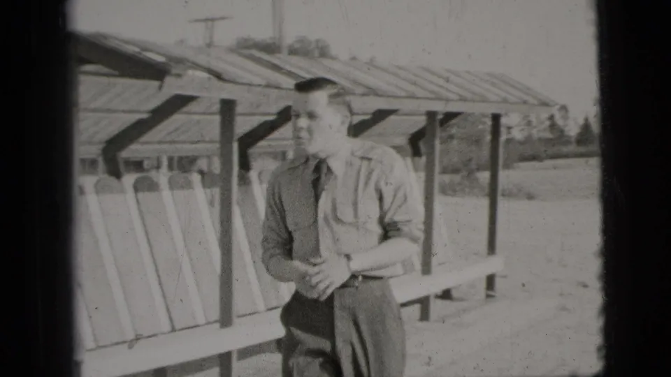 The man turns toward the camera near the wooden slatted shelter, with open ground and trees behind him.