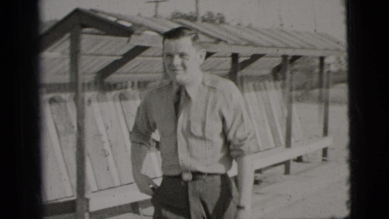 Young man in shirt, tie, and grey trousers stands beside a wooden shelter.