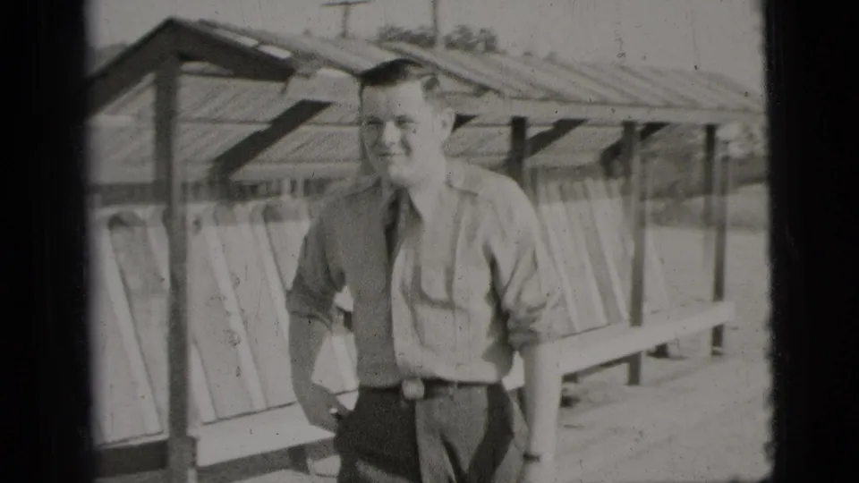 In 1956 New York City home-movie footage, a young man in a light shirt, dark tie, and grey trousers stands near a wooden shelter.