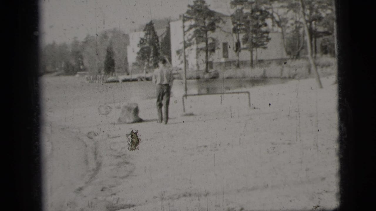 1956 New York City home-movie frame showing a man from behind near water, trees, a low goal frame, and a light building.