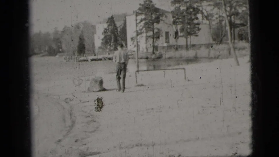 1956 New York City home-movie frame showing a man from behind near water, trees, a low goal frame, and a light building.