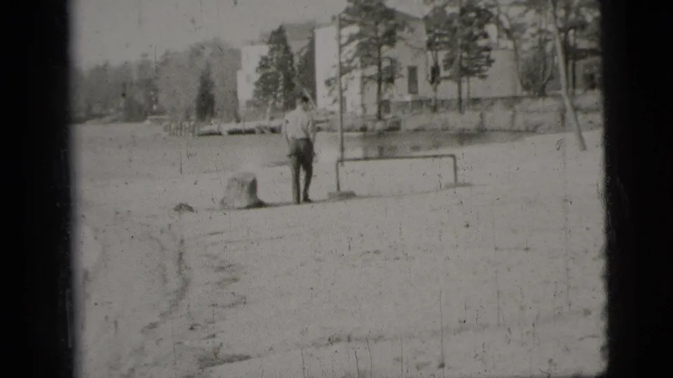 Black-and-white 8mm view of a man in a light shirt and dark pants near a low goal frame, trees, and water in New York City.