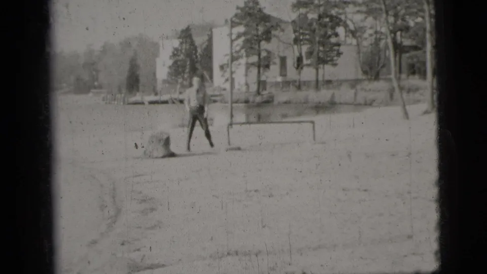 1956 New York City frame of a man walking across a sandy or gravelly area beside water, with trees and a building in the background.
