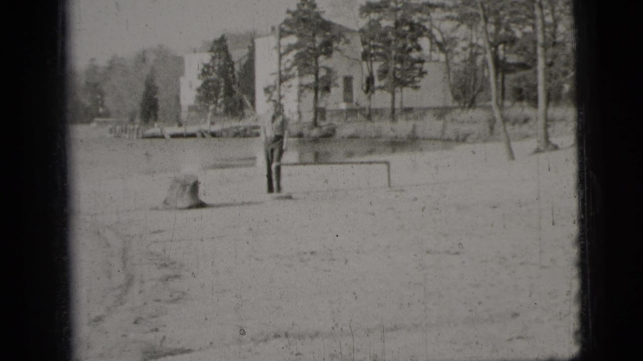 Man stands near a small goalpost in a sandy outdoor area with trees and buildings behind him.