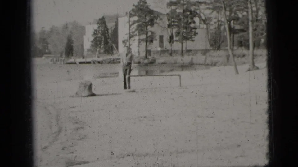 1956 New York City home-movie frame showing a man near a small goalpost on sandy ground with trees and buildings in the background.