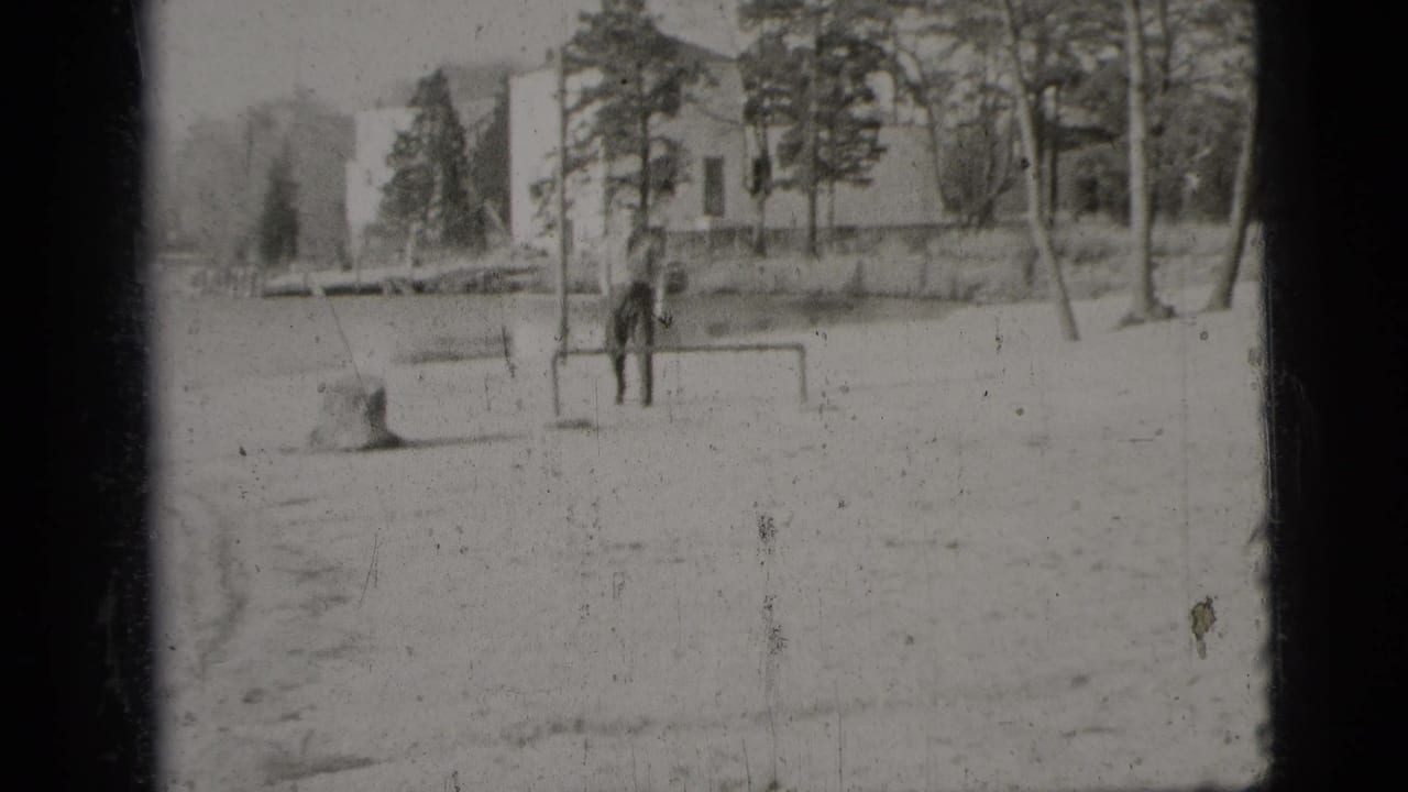 A man stands beside a small goalpost in an open sandy area, with trees and buildings beyond.
