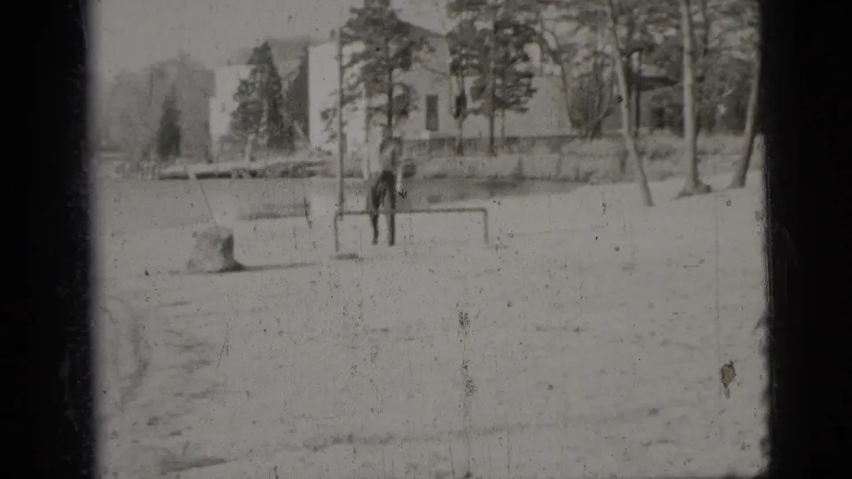 1956 New York City frame with a man near a low goalpost in a sandy outdoor space, backed by trees and light-colored buildings.