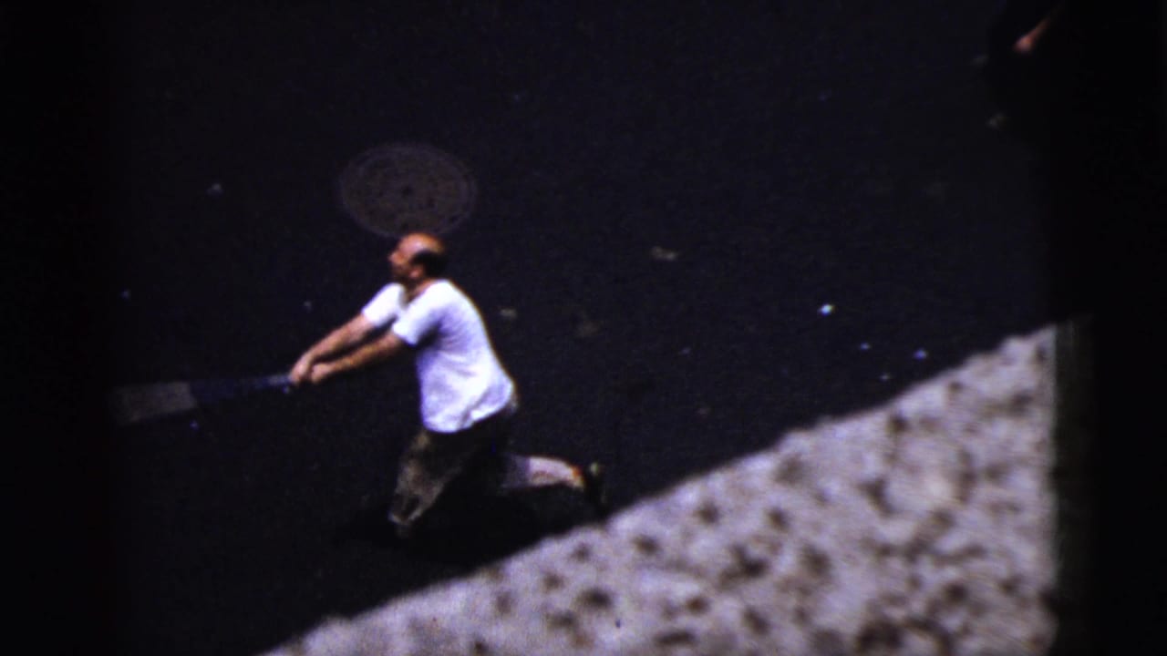 High-angle 1956 New York City street view of an adult man in a white shirt swinging a bat on dark pavement.